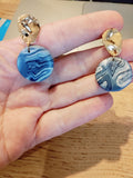 Paar ronde blauw gemarmerde oorbellen met goudkleurige stekers op een hand